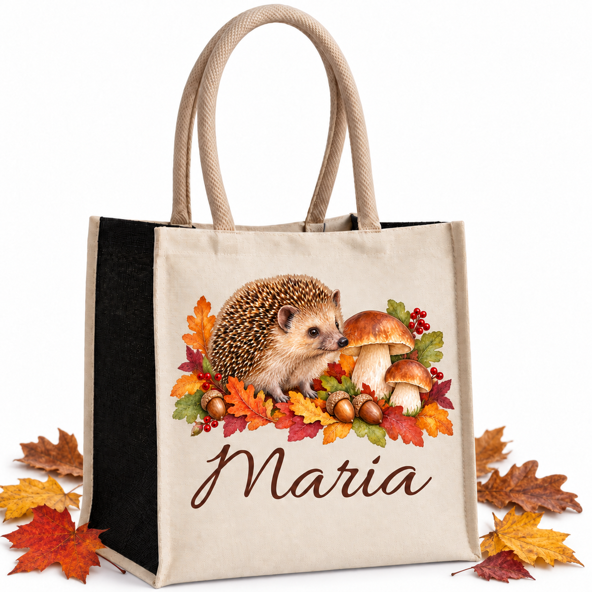 Baumwoll-Jute-Tasche mit Igel und Pilzen und Wunschname, naturfarbene Front und schwarze Seiten, mit echten Herbstblättern neben der Tasche auf hellem Hintergrund.