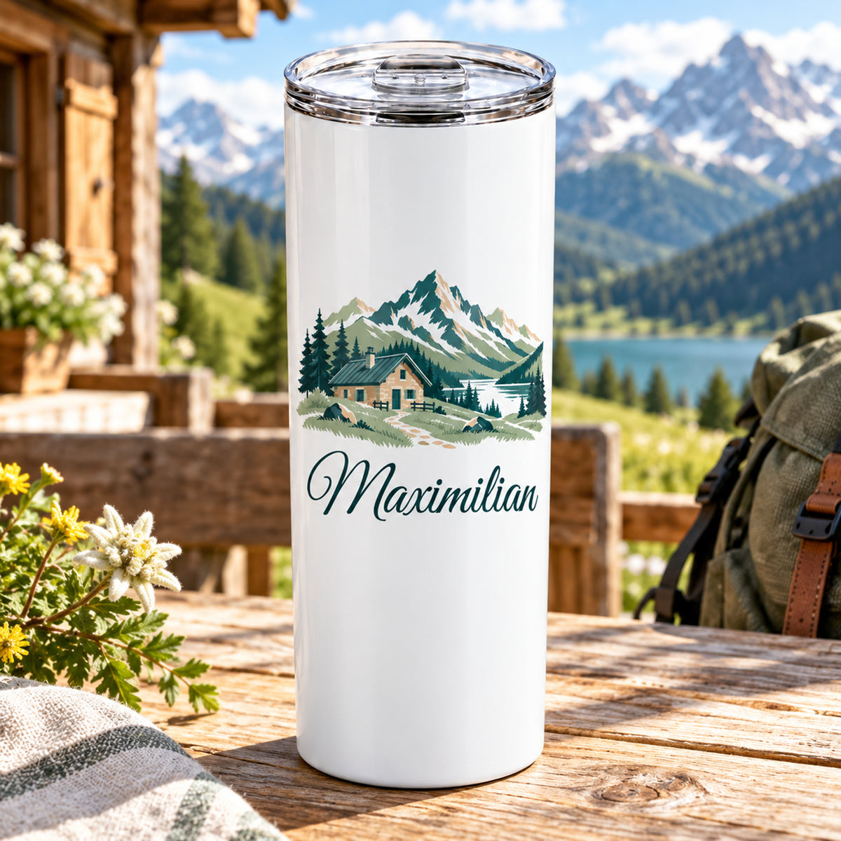 Thermobecher mit Berghütte und Bergen und Wunschname auf einem Tisch vor einer Berghütte.