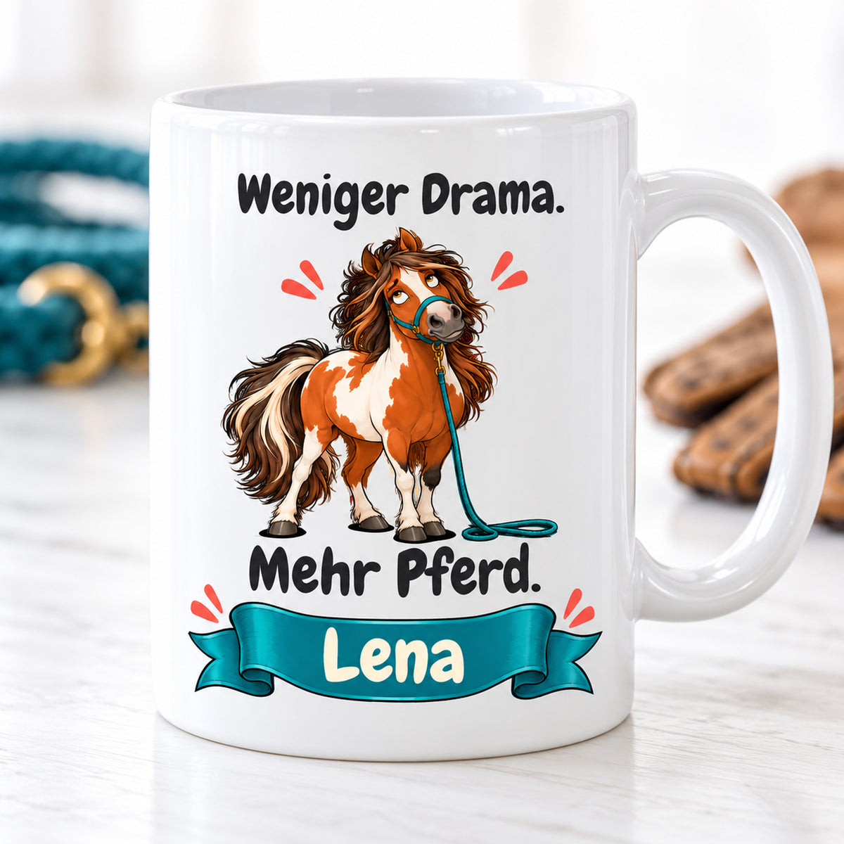 Weiße Pferdetasse mit Namen, Schecken mit petrolfarbenem Strick und Spruch „Weniger Drama. Mehr Pferd.“, auf heller Ablage.