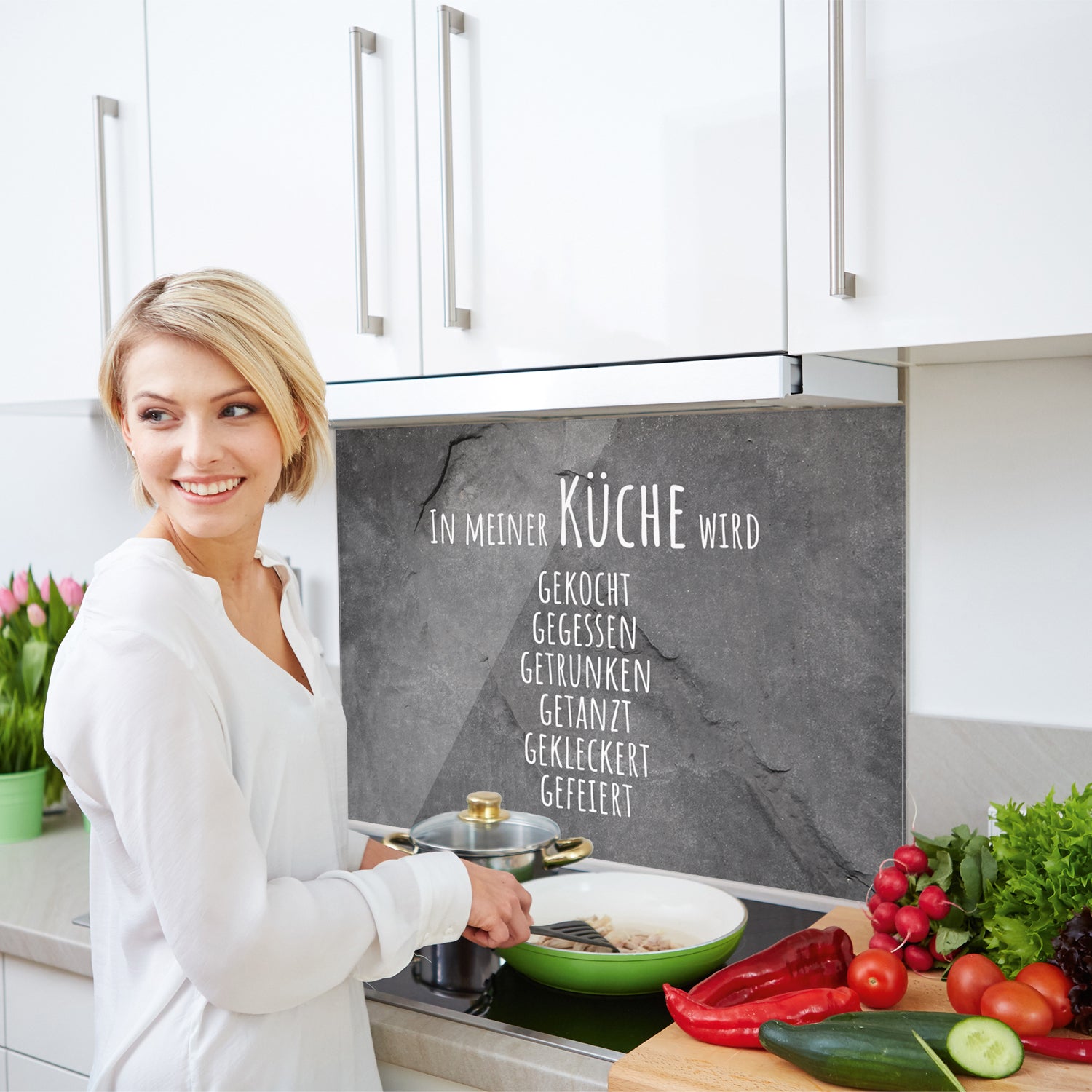 Küchenwand mit Schriftzug, Frau kocht mit frischem Gemüse.