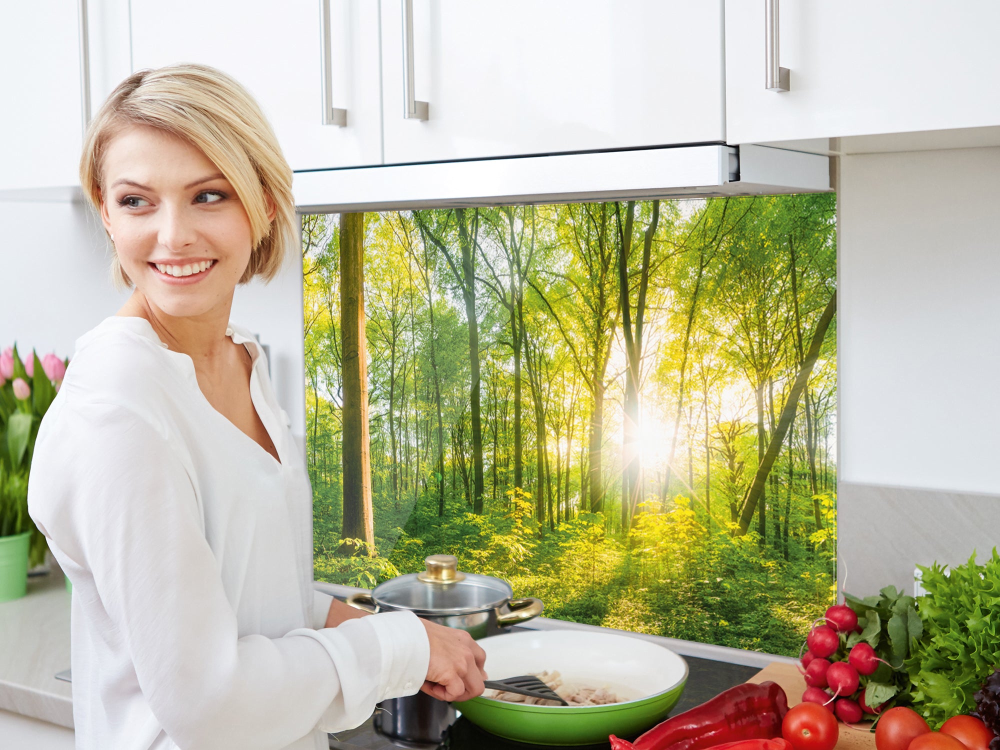 Frau mit Pfanne in heller Küche, Waldmotiv im Hintergrund.