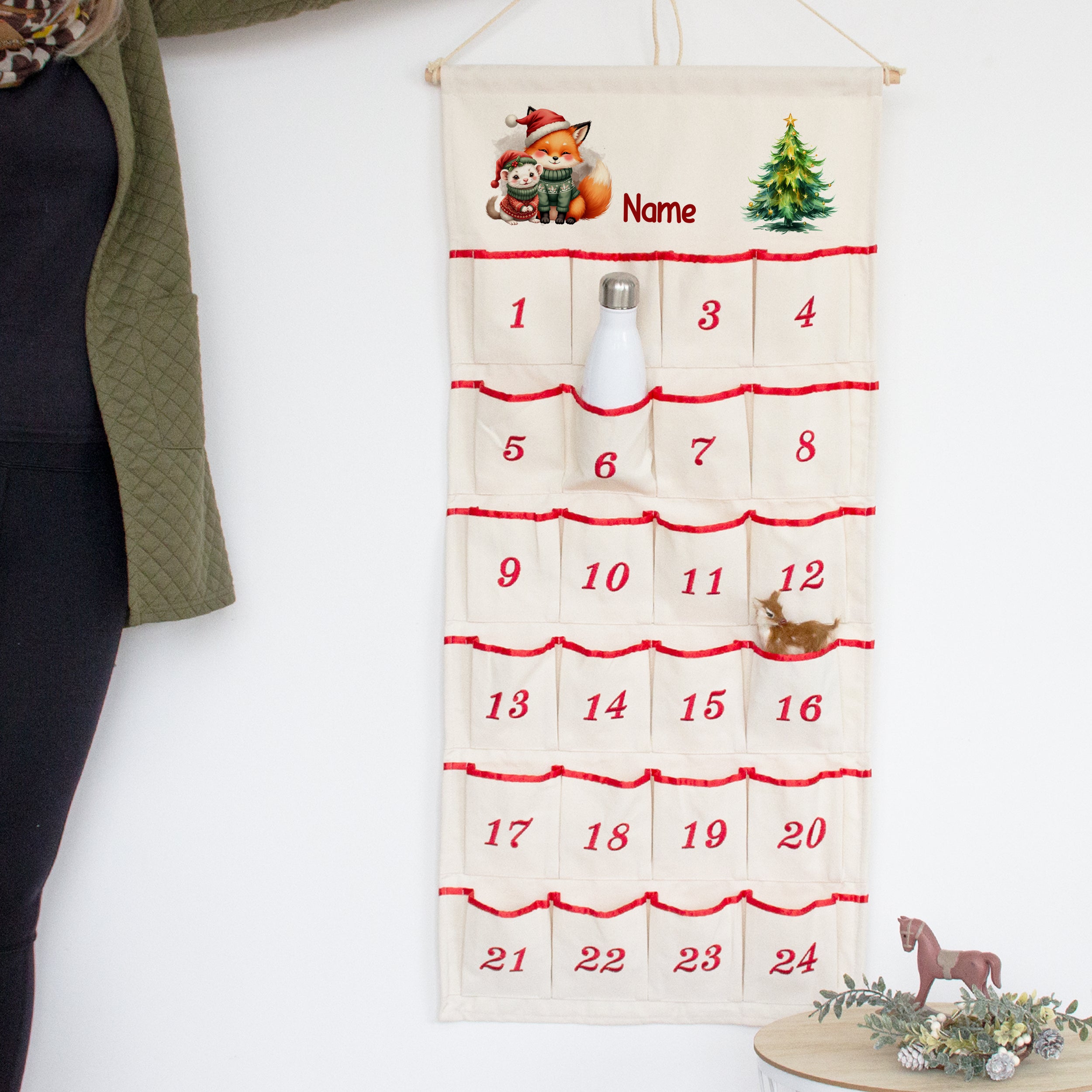 Adventskalender mit Wunschname, Fuchs und kleinem Waldtier, an der Wand mit Flasche in einer nummerierten Tasche